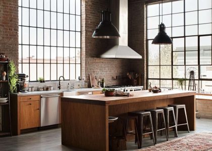 Loft kitchen with exposed brick walls, polished concrete floors, metal light fixtures, rustic wooden island, large windows letting in natural light, aged cabinetry, and a vintage rug, creating an inviting and cozy culinary space.
