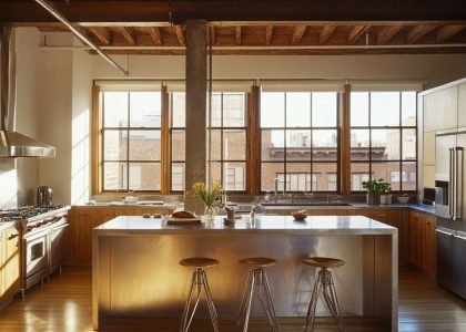 Loft kitchen with high ceilings, exposed wooden beams, a metal island, sleek cabinetry, warm wood accents, large windows allowing sunlight, and a cozy atmosphere, perfect for gatherings.